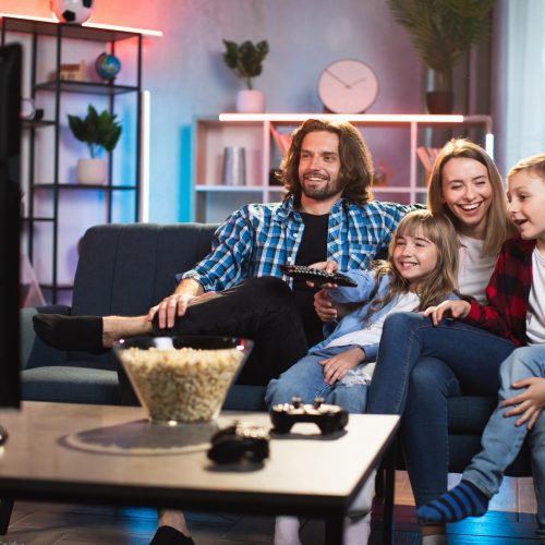 Smiling young parents with two kids sitting on couch and watching TV. Happy caucasian family in casual wear spending evening time together at home.